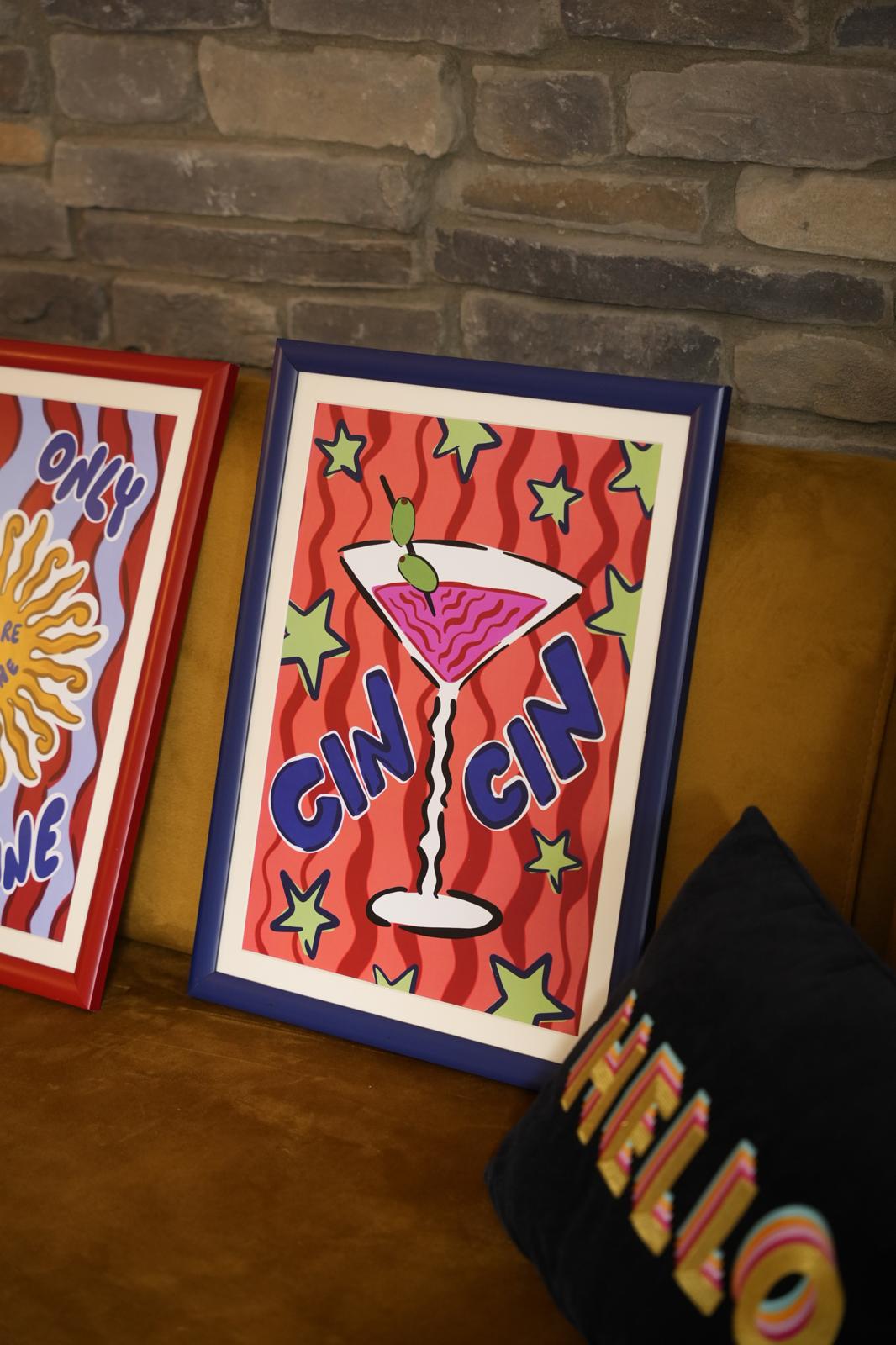 Framed Cin Cin cocktail print beside a colourful sunshine print on a mustard sofa.