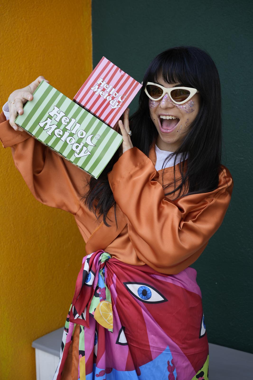Woman holding pink and green striped Hello Melody gift boxes against a colourful wall.