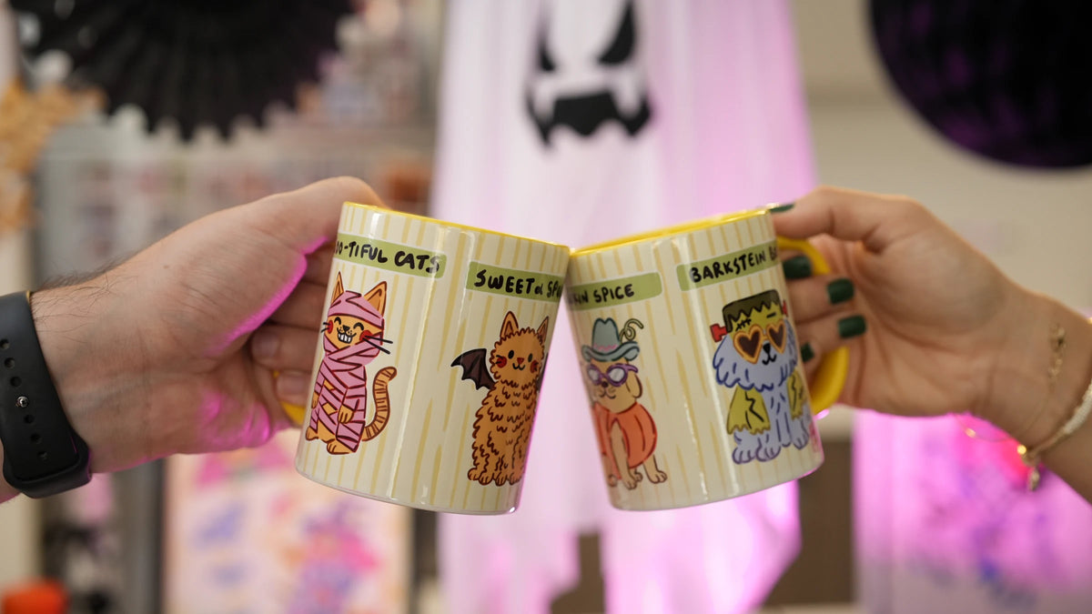 Two hands clinking Hello Melody Halloween mugs, Boo-tiful Cats and Barkstein Boo, in front of a pink ghost decoration